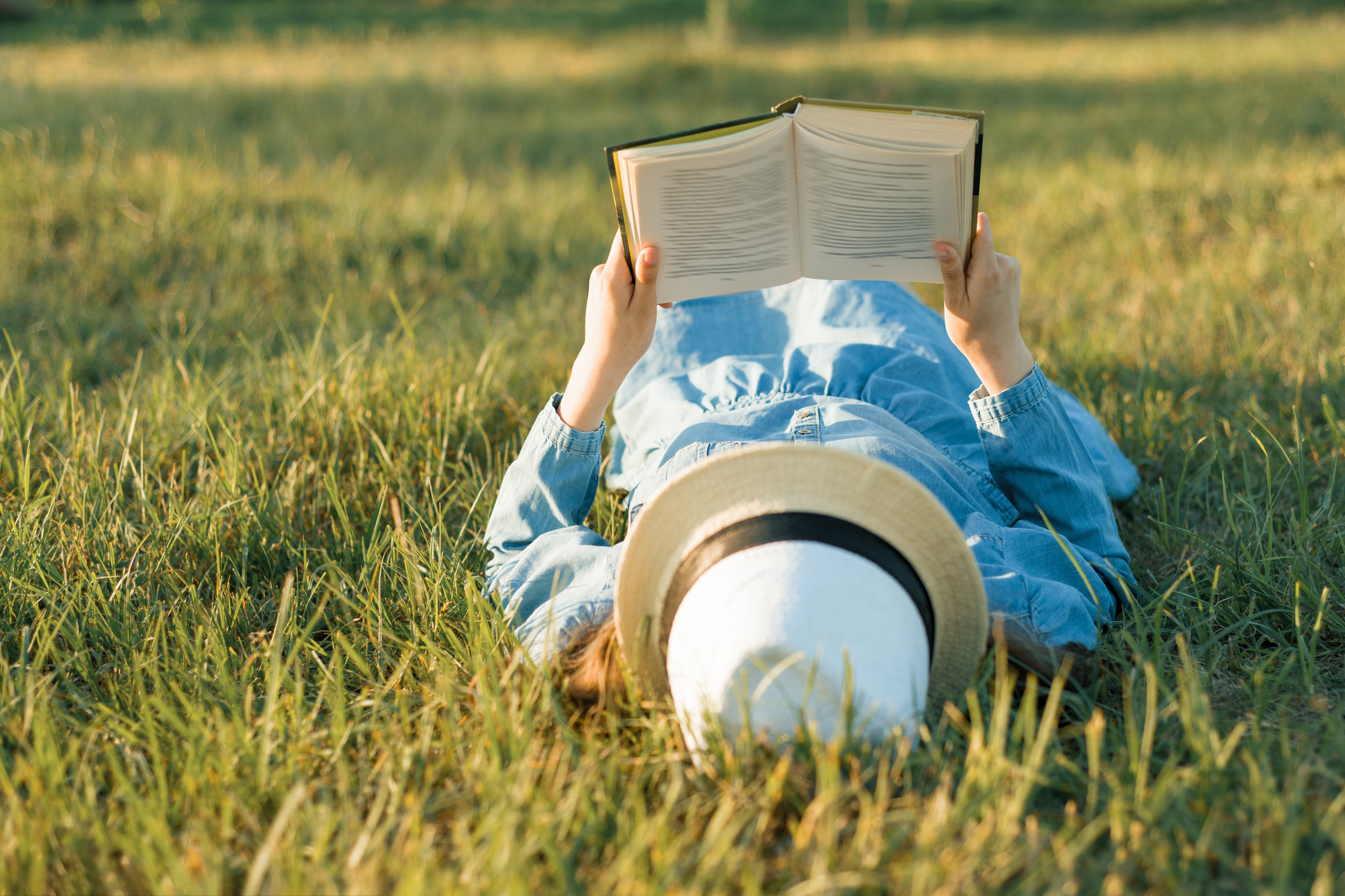 Girl in dress and hat lies on green grass reading book. View from above.